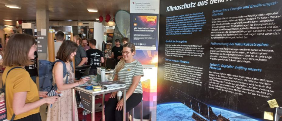 Ein Messestand, der über Erdbeobachtung und Weltraumaktivitäten in Österreich informiert. Davor stehen interessierte Besucher:innen, die aufmerksam die ausgestellten Plakate studieren.