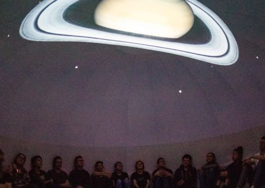Kinder sitzenin einem abgedunkelten Raum am Boden über ihnen ist der Saturn auf die Decke projiziert.