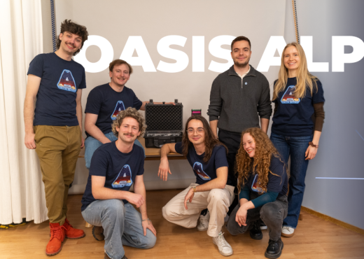 4 junge Männer und 2 Frauen mit dunkelblauen Oasis Alpha T-Shirt und ein junger Mann im grauen Pullover lächeln strahlend in die Kamera. In ihrer Mitte ist ein kleiner gepolsterter schwarzer Koffer, hinter ihnen der Schriftzuug Oasis Alpha und rechts von ihnen ein 3D Modell ihres Satelliten zu sehen.